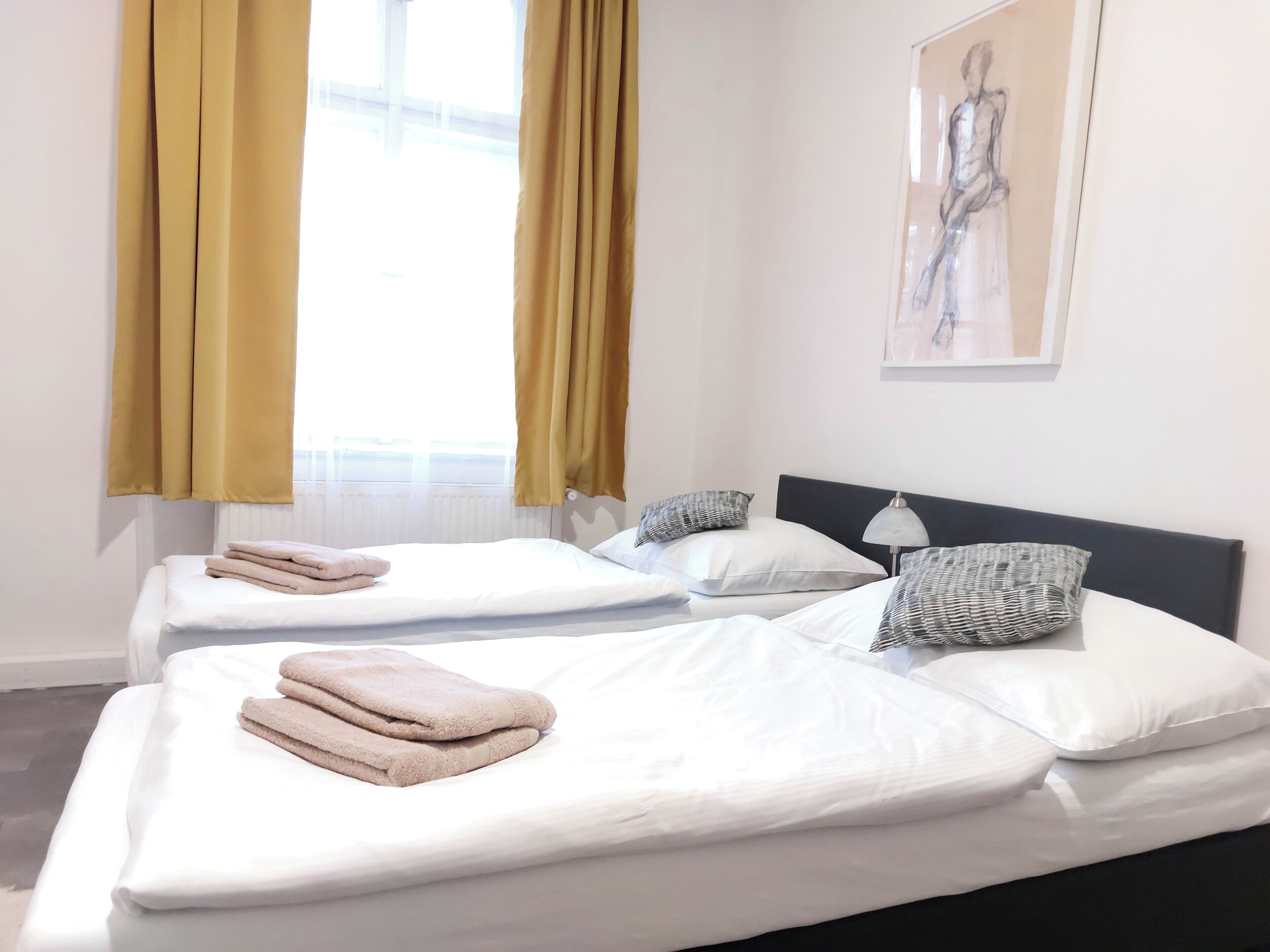 A hotel room with two beds, each with white linens and a folded towel on the left. There are patterned pillows and a lamp on the right side of the beds. A window with yellow curtains is visible, and a painting hangs on the wall above the beds.