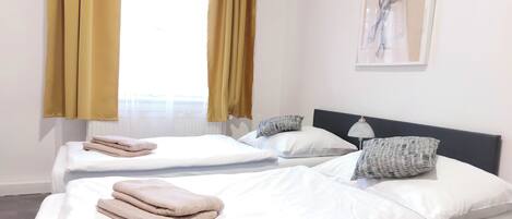 A hotel room with two beds, each with white linens and a folded towel on the left. There are patterned pillows and a lamp on the right side of the beds. A window with yellow curtains is visible, and a painting hangs on the wall above the beds.