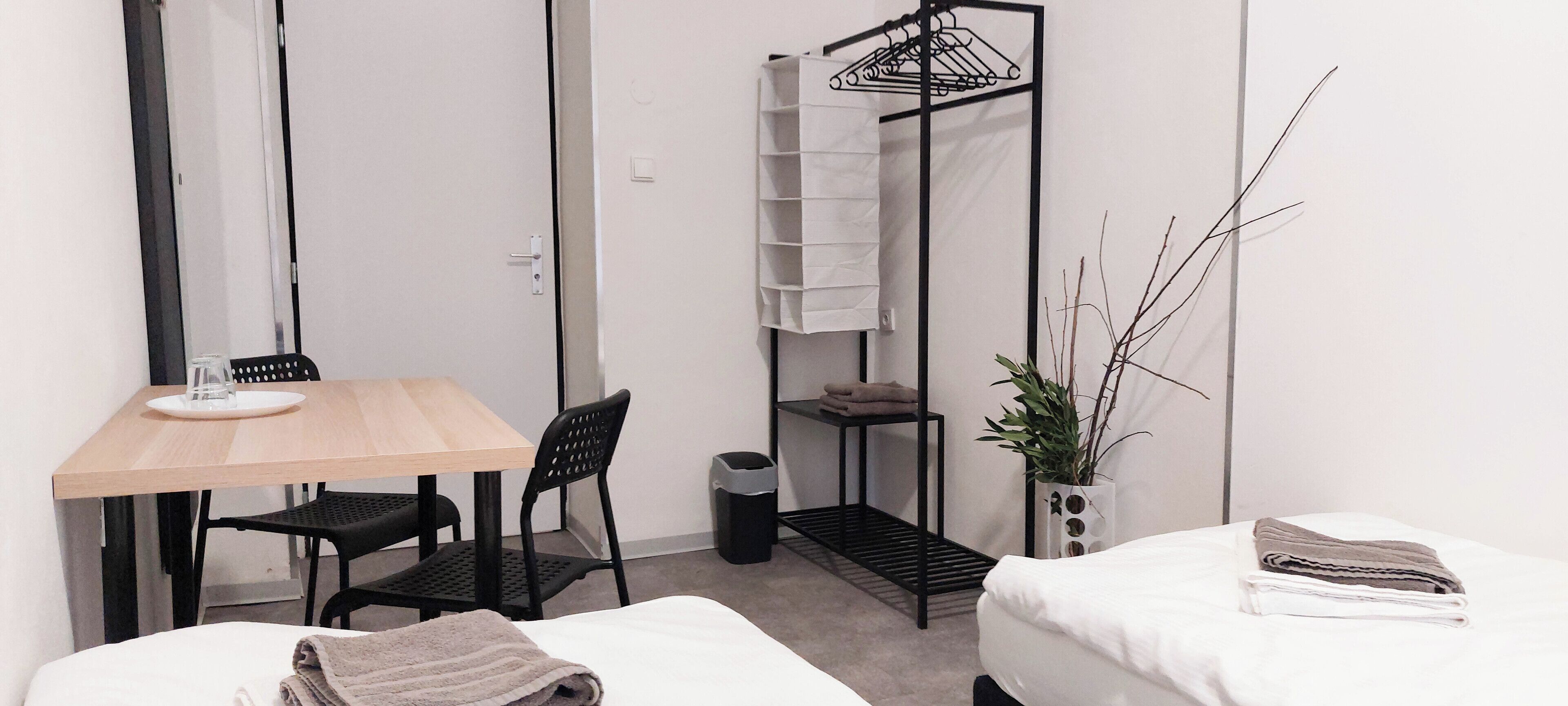 A small, clean room with a wooden table, two black chairs, a bed with a white comforter, a small shelf with folded towels, a potted plant, and a black hanging rack with folded clothes.