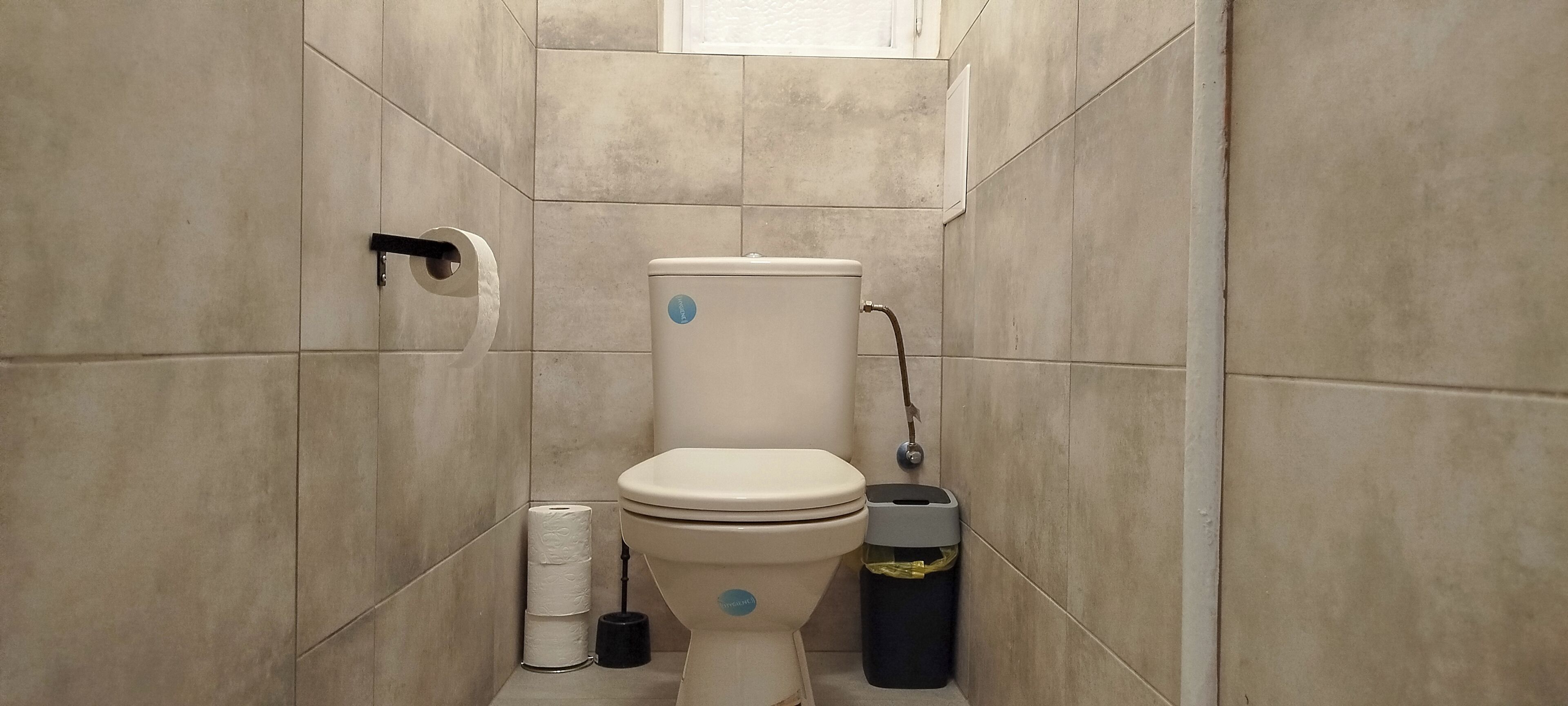 A bathroom with a toilet, a roll of toilet paper, a trash bin, and a wall-mounted toilet paper holder.