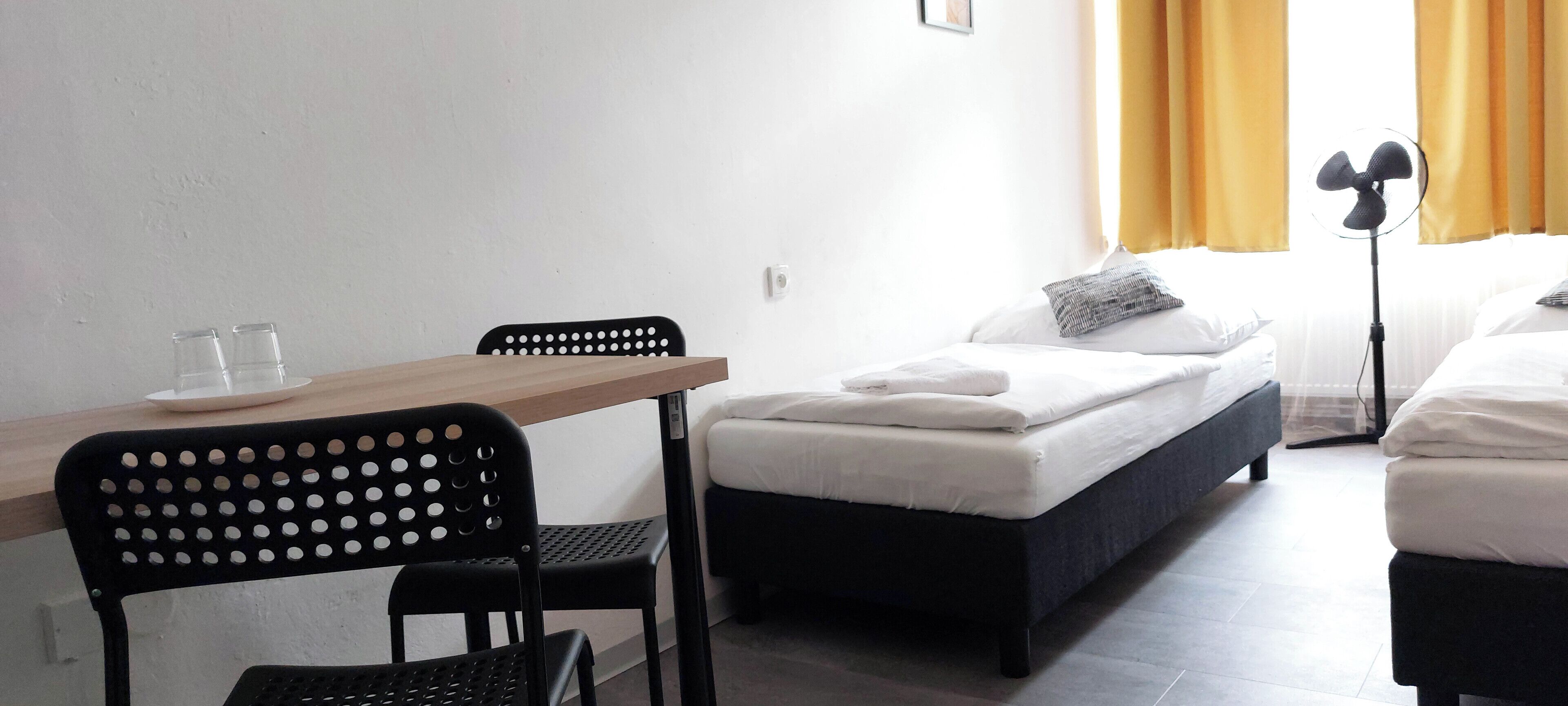 Ein Hotelzimmer mit einem Einzelbett, einem kleinen Tisch mit zwei Gläsern, einem Stuhl und einem Standventilator.