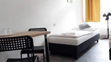 Ein Hotelzimmer mit einem Einzelbett, einem kleinen Tisch mit zwei Gläsern, einem Stuhl und einem Standventilator.