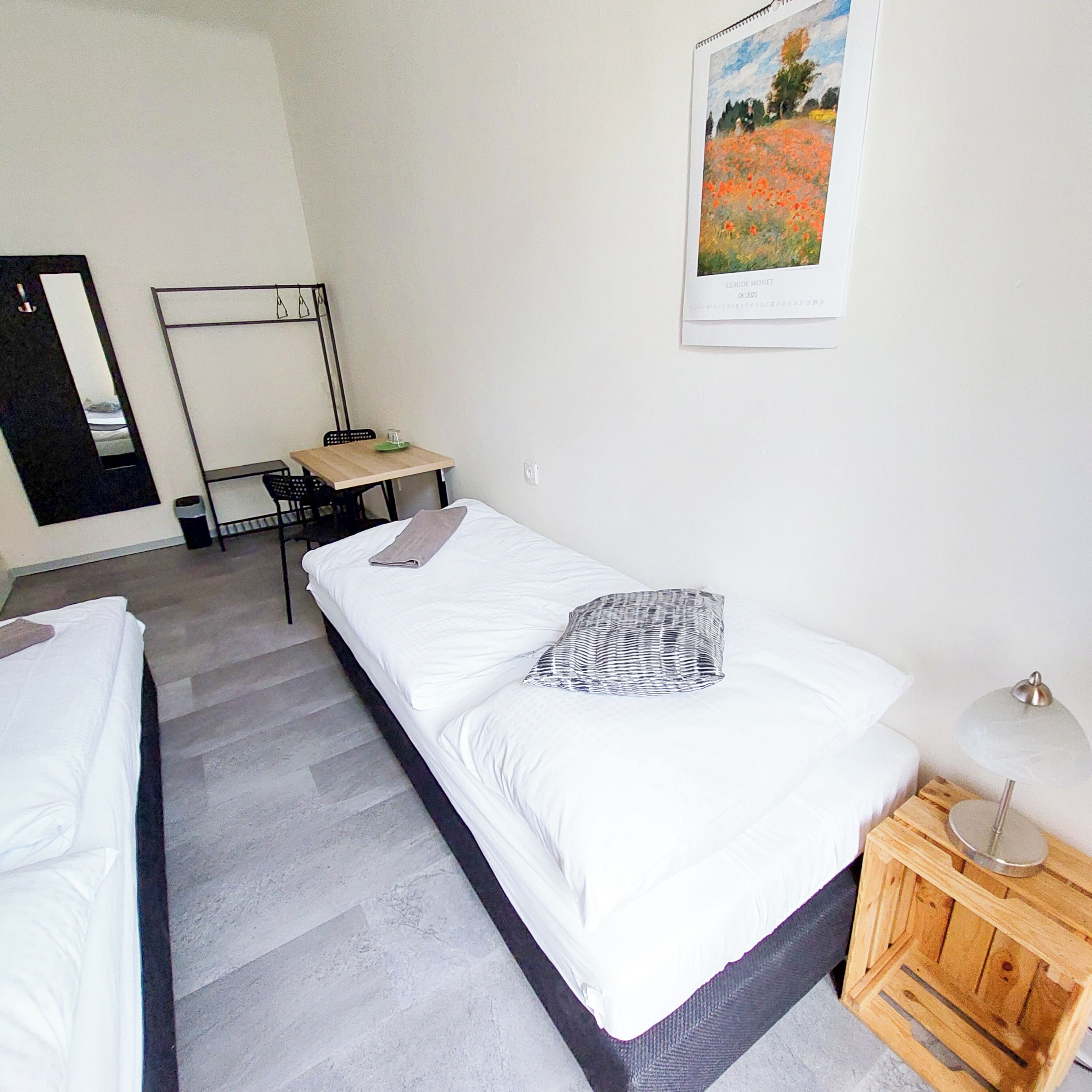 A room with two single beds, a desk, a chair, a small table, a lamp, and a poster on the wall.