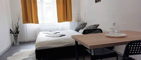 A hotel room with a bed, a table with a tray, a chair, a potted plant, and a window with curtains.
