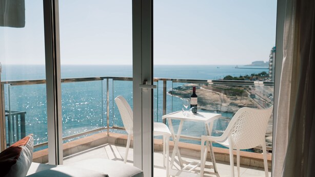 A balcony with a table and chairs, overlooking a coastal view with a clear sky.