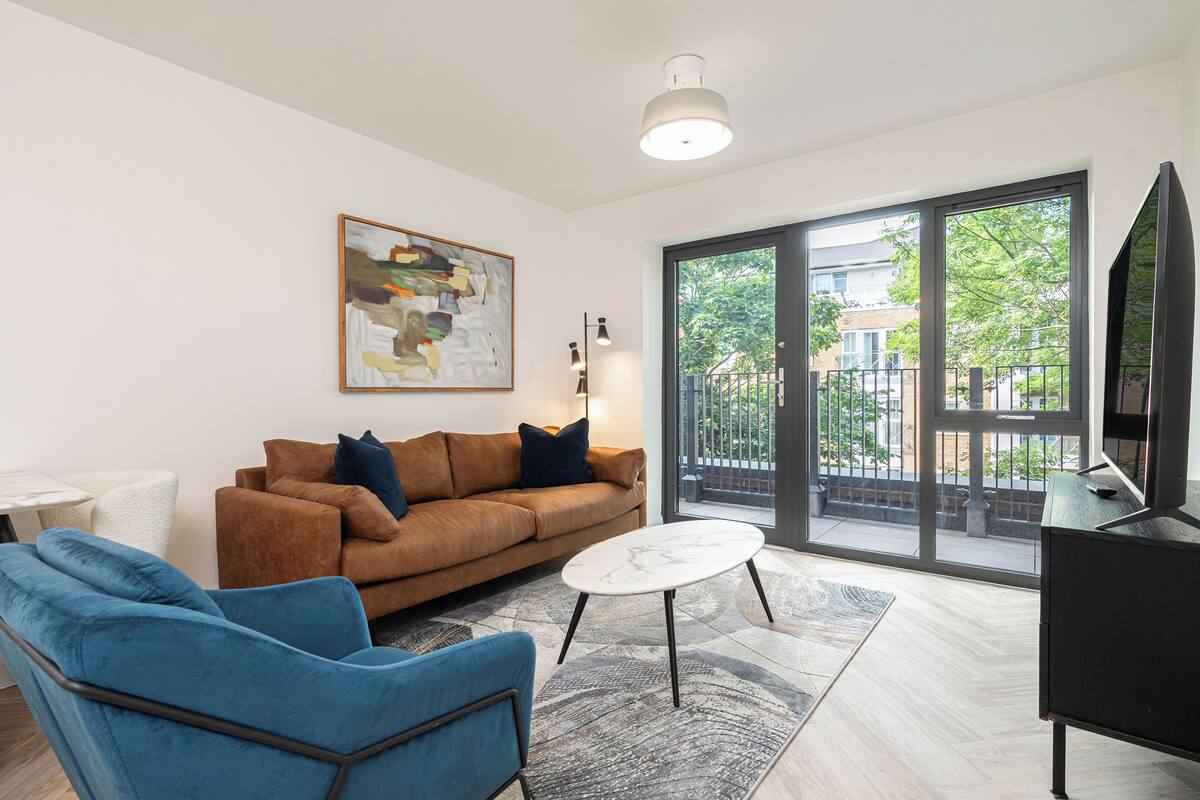 Una sala de estar moderna con un sofá marrón, un sillón azul y una mesa de centro blanca. Un cuadro cuelga de la pared, y hay un ventanal con vistas a edificios y árboles.
