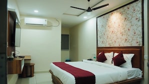 A hotel room with a bed, a ceiling fan, an air conditioning unit, a wooden bedside table, a small stool, and a wall-mounted television.