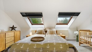 Ein Schlafzimmer mit schräg verlaufender Decke, zwei Dachfenstern, einem Bett mit einem Anker-Muster-Überwurf, einem hölzernen Kinderbett und einer Sterndeko.