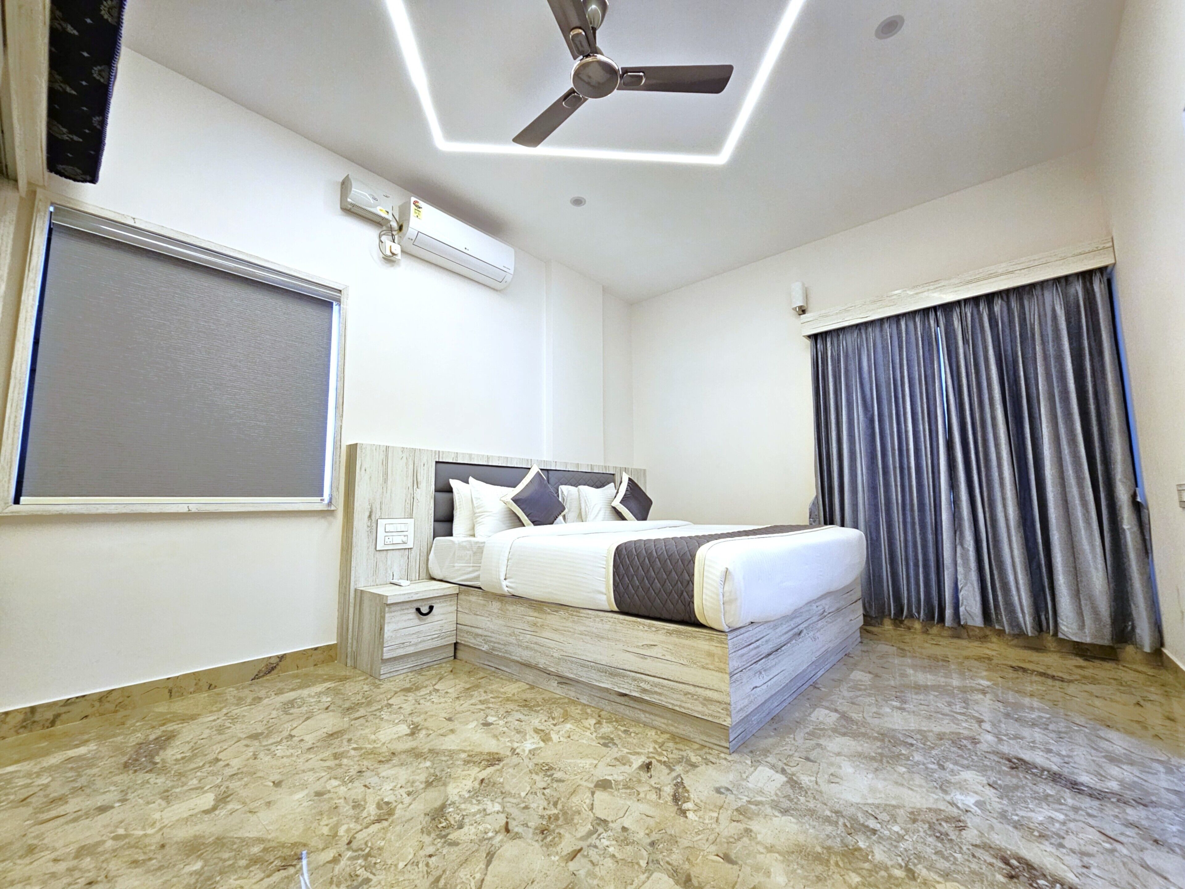 A modern hotel room with a bed, bedside tables, a ceiling fan, an air conditioning unit, and a large window with curtains.