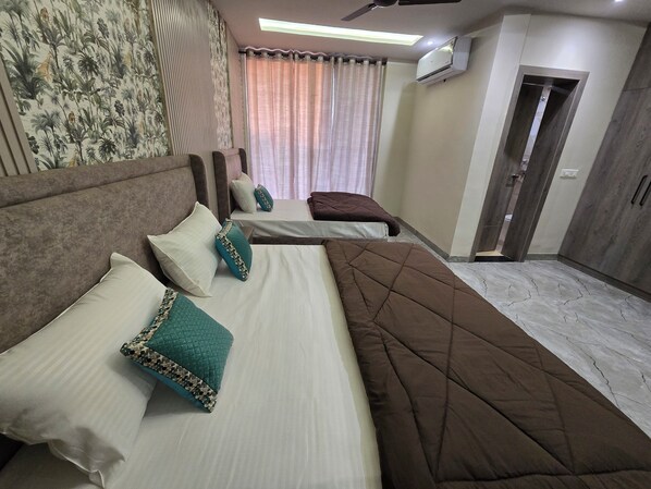 A hotel room with a large bed, two smaller beds, a ceiling fan, an air conditioning unit, and a door leading to another room.