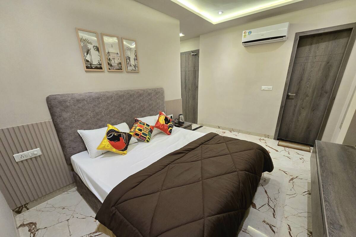 Habitación de hotel con cama, cabecera gris, tres almohadas decorativas, colcha acolchada marrón, aire acondicionado de pared y puerta con marco de madera.