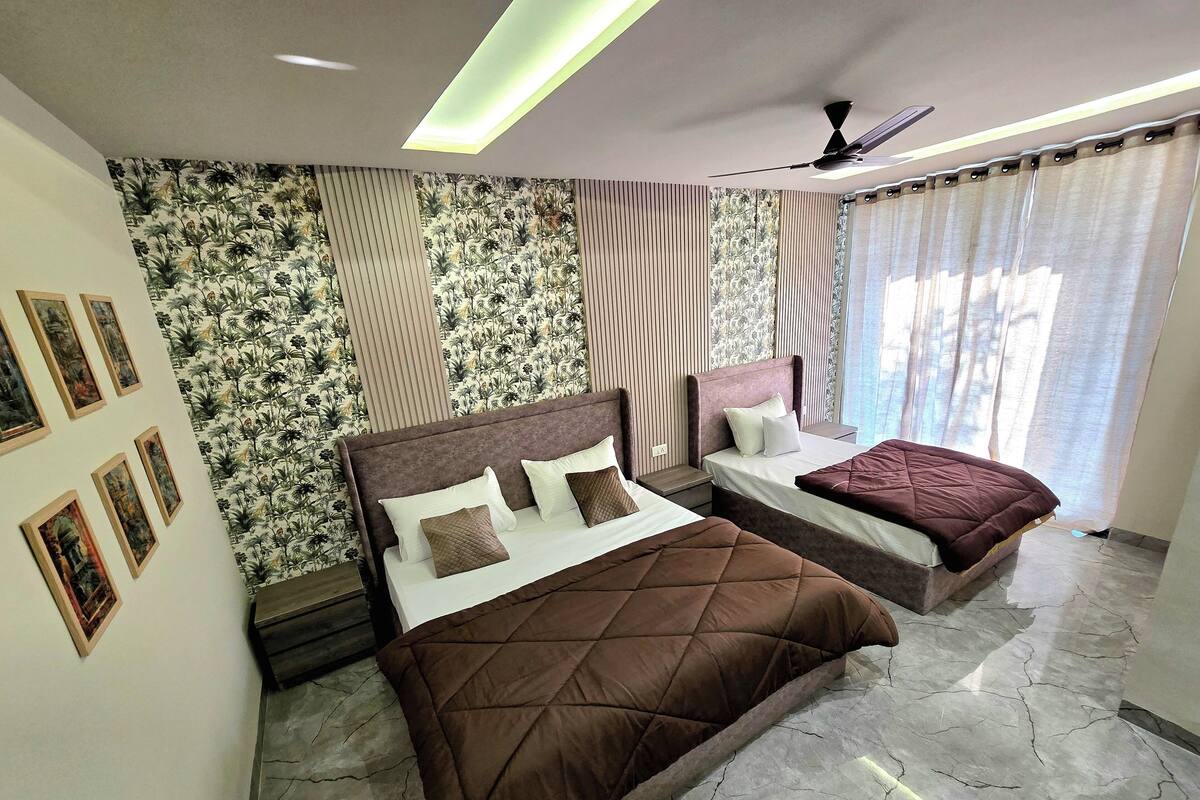 Un dormitorio con una cama grande, una cama más pequeña y un sofá. Hay varias sábanas y almohadas. La habitación tiene un ventilador de techo y una ventana con cortinas.