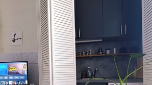 A modern kitchen with dark cabinets, a built-in stove, and a small refrigerator. There is a television on a stand displaying a movie, and a potted plant in the corner.