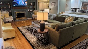 A modern living room with a green sofa, a coffee table, and a TV displaying a show. There is a fireplace, a dining area, and a kitchen in the background. - Cherry Tree Vacation Home, 4 BR 2 BA 2500 sq ft, safe neighborhood .9 mi to ND! (South Bend)