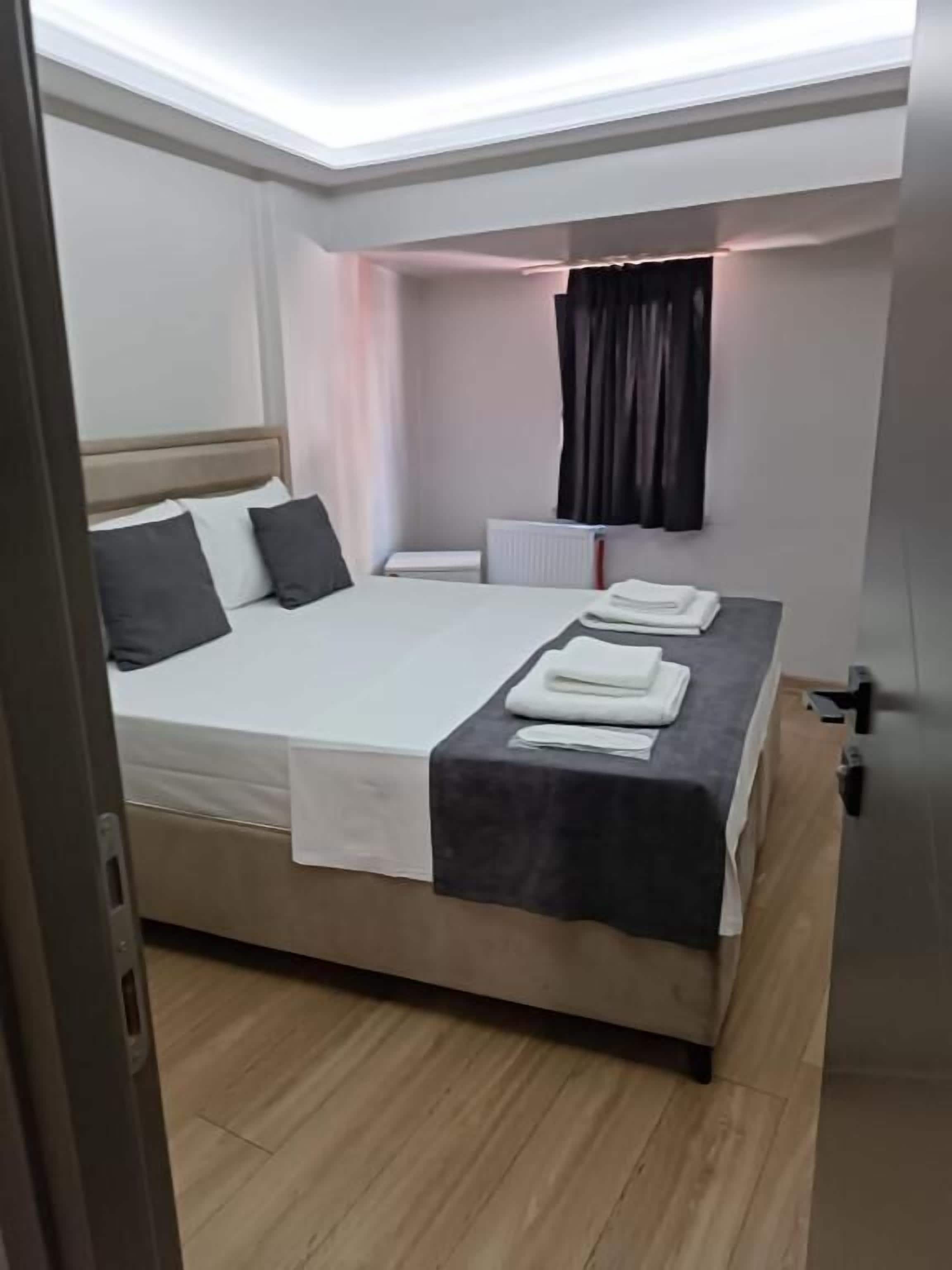 Ein ordentlich bezogenes Bett mit weißen Laken und einer dunklen Decke, ein Nachttisch mit einem Buch und einer Lampe sowie ein Fenster mit Vorhängen.