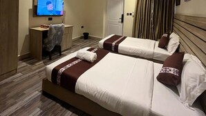 A hotel room with two beds, a desk, a television, and a door. - Moz One Apartments (Abuja)