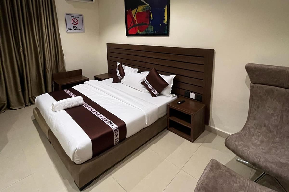 A hotel room with a large bed, a wooden headboard, a nightstand, an air conditioning unit, a painting, a "No Smoking" sign, and a chair.
