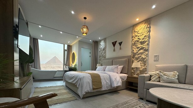 A modern bedroom with a large bed, a sofa, a desk, and a view of a pyramid. - First Row to Pyramids Studio (Giza Governorate)