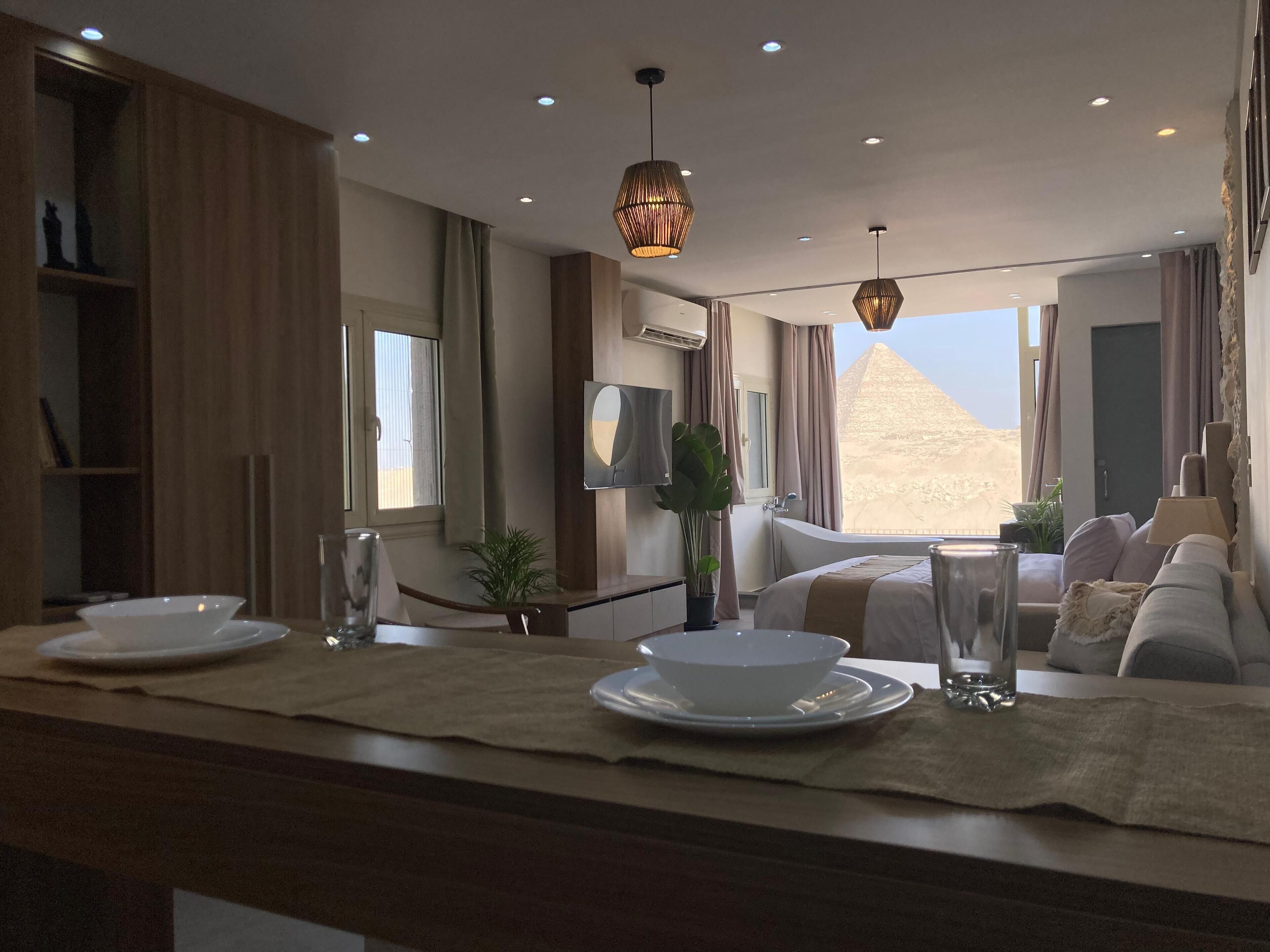 A modern hotel room with a large window, a bathtub, a sofa, and a dining area with plates and glasses.