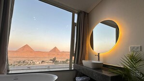 A modern bathroom with a bathtub, a sink, and a large mirror. A view of the pyramids is visible through the window. - First Row to Pyramids Studio (Giza Governorate)