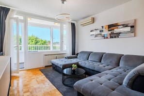 A modern living room with a grey sectional sofa, a round black coffee table, and a large window with a view of greenery.