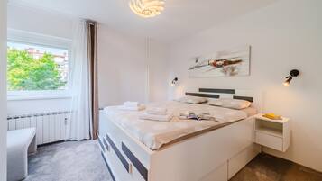 A modern bedroom with a large bed, a bedside table, a window with curtains, and a wall-mounted artwork.