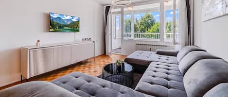 Een moderne woonkamer met een grote flatscreen televisie, een bank, een salontafel en uitzicht op groen door grote ramen.