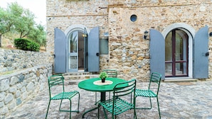 Edificio de piedra con una mesa redonda y cuatro sillas, una planta pequeña sobre la mesa, y dos ventanas arqueadas con contraventanas.