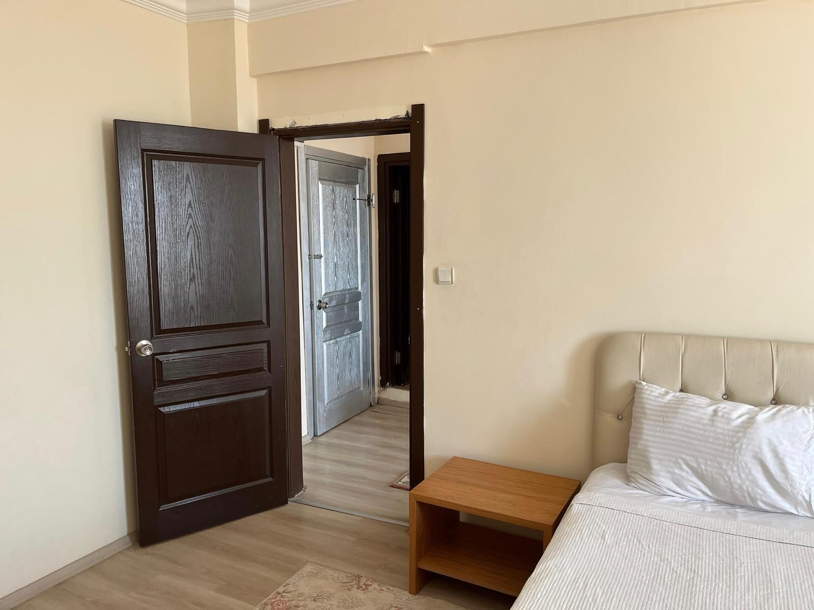 A bedroom with a bed, a wooden nightstand, a dark wooden door, and a hallway leading to another room.