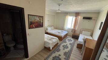 Une chambre d’hôtel avec deux lits, un bureau, une chaise, une petite table, un tableau au mur, une fenêtre avec des rideaux et un ventilateur de plafond.