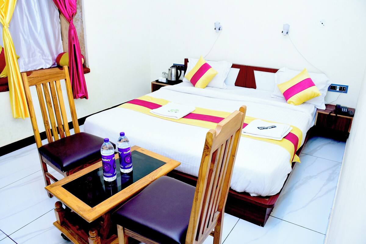 A hotel room with a bed, a wooden chair, a small table with a chair, and two bottles of water.