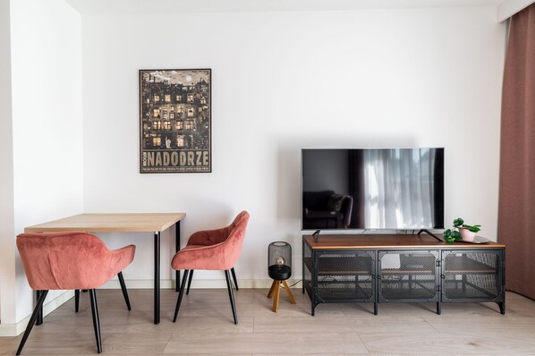 A modern living room with a wooden table, two pink chairs, a black TV, and a decorative poster on the wall. - RentPlanet - Apartament Ptasia (Wrocław)