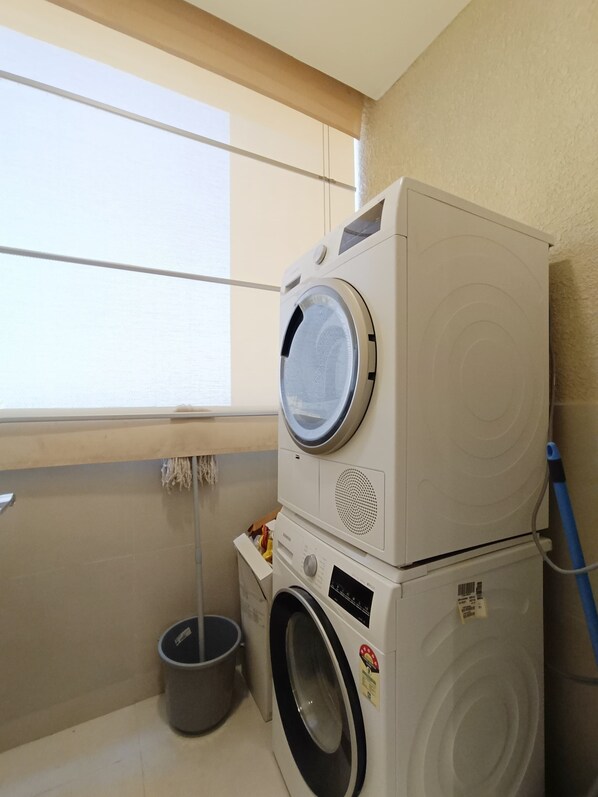 A compact laundry area with stacked washing machines, a small trash bin, and a window for natural light.