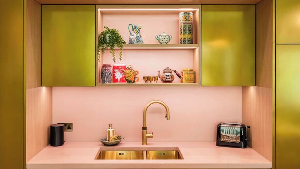 A modern kitchen with a pink countertop, gold fixtures, and a shelf with decorative items.