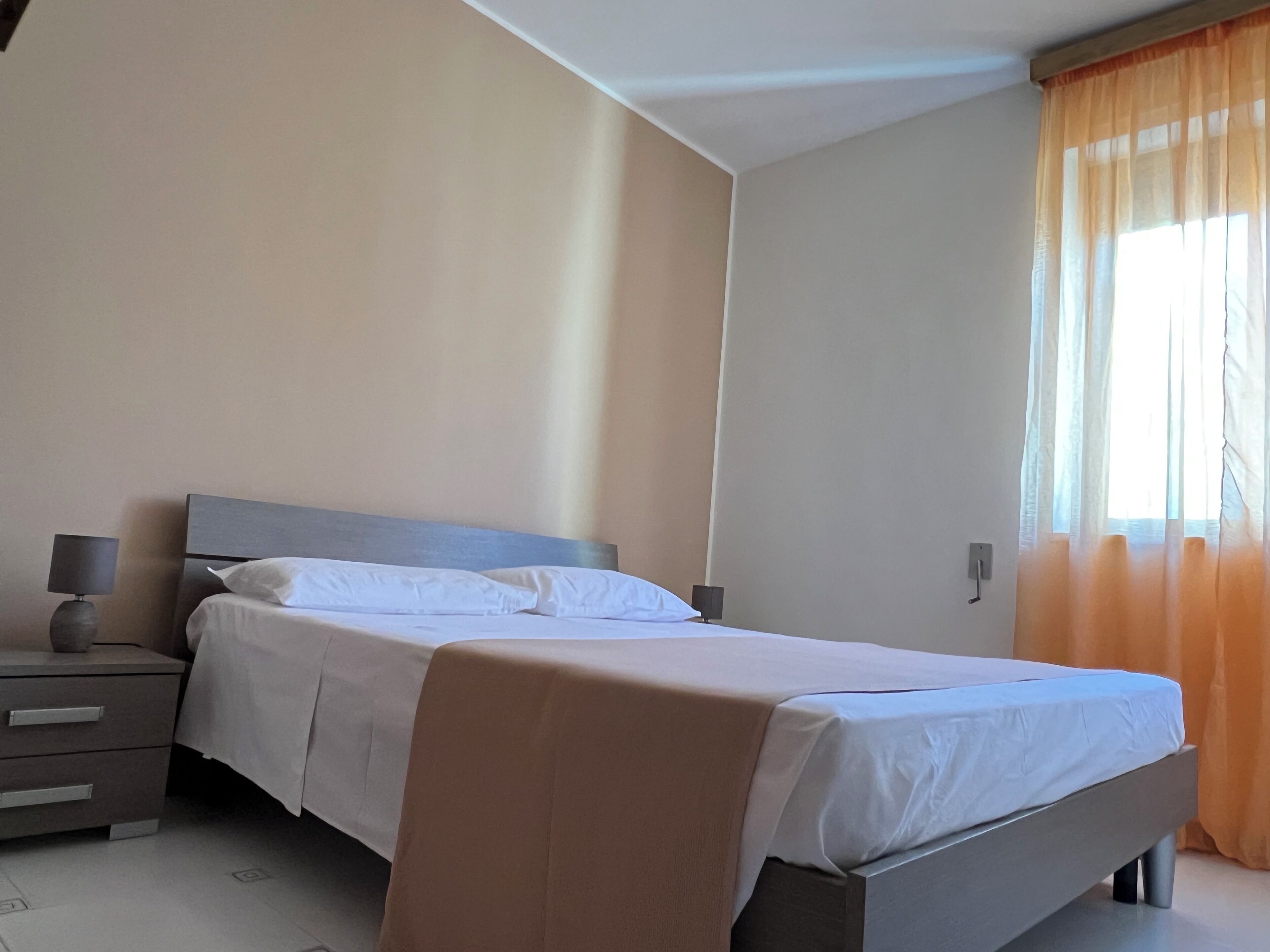 Una cama bien hecha en una habitación con una mesita de noche, una lámpara y una ventana con cortinas.