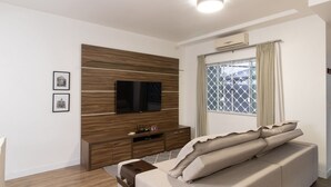 Un salon moderne avec un téléviseur à écran plat fixé sur un mur recouvert de panneaux en bois, un canapé agrémenté de coussins décoratifs, une petite table basse en bois et une fenêtre avec un grillage métallique.