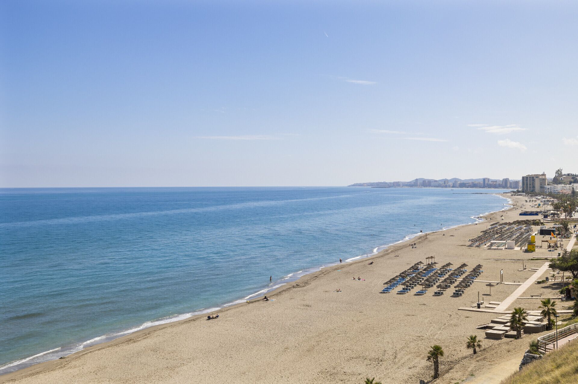 Ein Strand mit einer Reihe blauer Sonnenschirme und Liegestühlen, einem sandigen Ufer und einer Stadtansicht im Hintergrund.