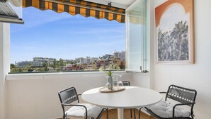 A balcony with a white table, two wicker chairs, a vase with flowers, and a cityscape view. - Fuengirola Sea View Apartment–Pool, Tennis & Beach (Benalmádena)