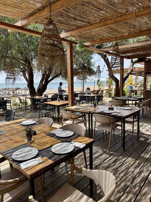 Un área de comedor al aire libre con mesas y sillas, una terraza de madera y vista a la playa.