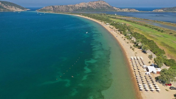Luchtbeeld van een kustgebied met een strand, helder water en een bergachtige achtergrond.