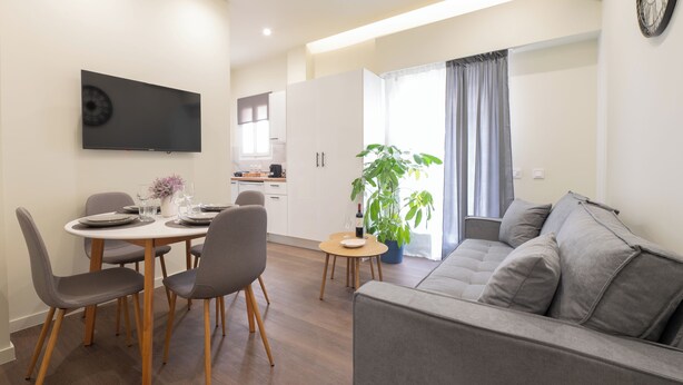 Un salon moderne avec un canapé gris, une table à manger avec des chaises, un téléviseur à écran plat fixé au mur et une plante en pot.