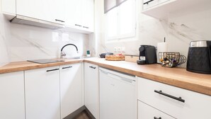Une cuisine moderne avec des armoires blanches, un plan de travail en bois, un lave-vaisselle encastré, une machine à café et un panier rempli d’ustensiles.