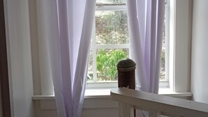 Een kamer met een raam, bedekt met witte gordijnen, een kleine potplant op de vensterbank en een witte balustrade op de voorgrond.