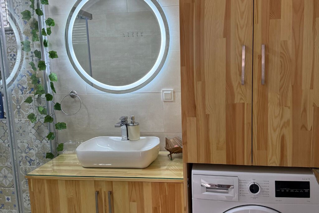 A bathroom with a wooden vanity, a white sink, a round mirror, and a washing machine.