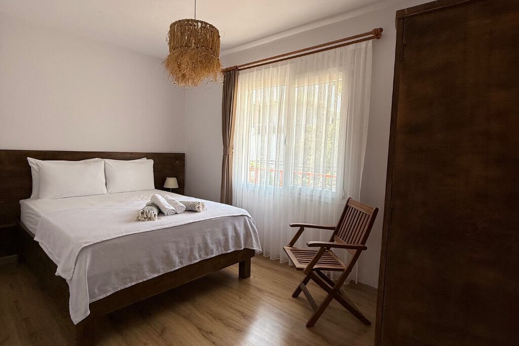 Una cama bien hecha con sábanas blancas, cabecera de madera, una silla de madera, lámpara colgante y un ventanal con cortinas traslúcidas.