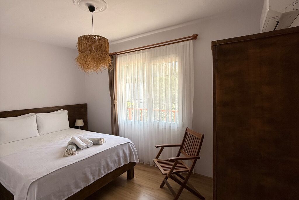 A bedroom with a bed, a chair, a hanging light fixture, and a window with sheer curtains.