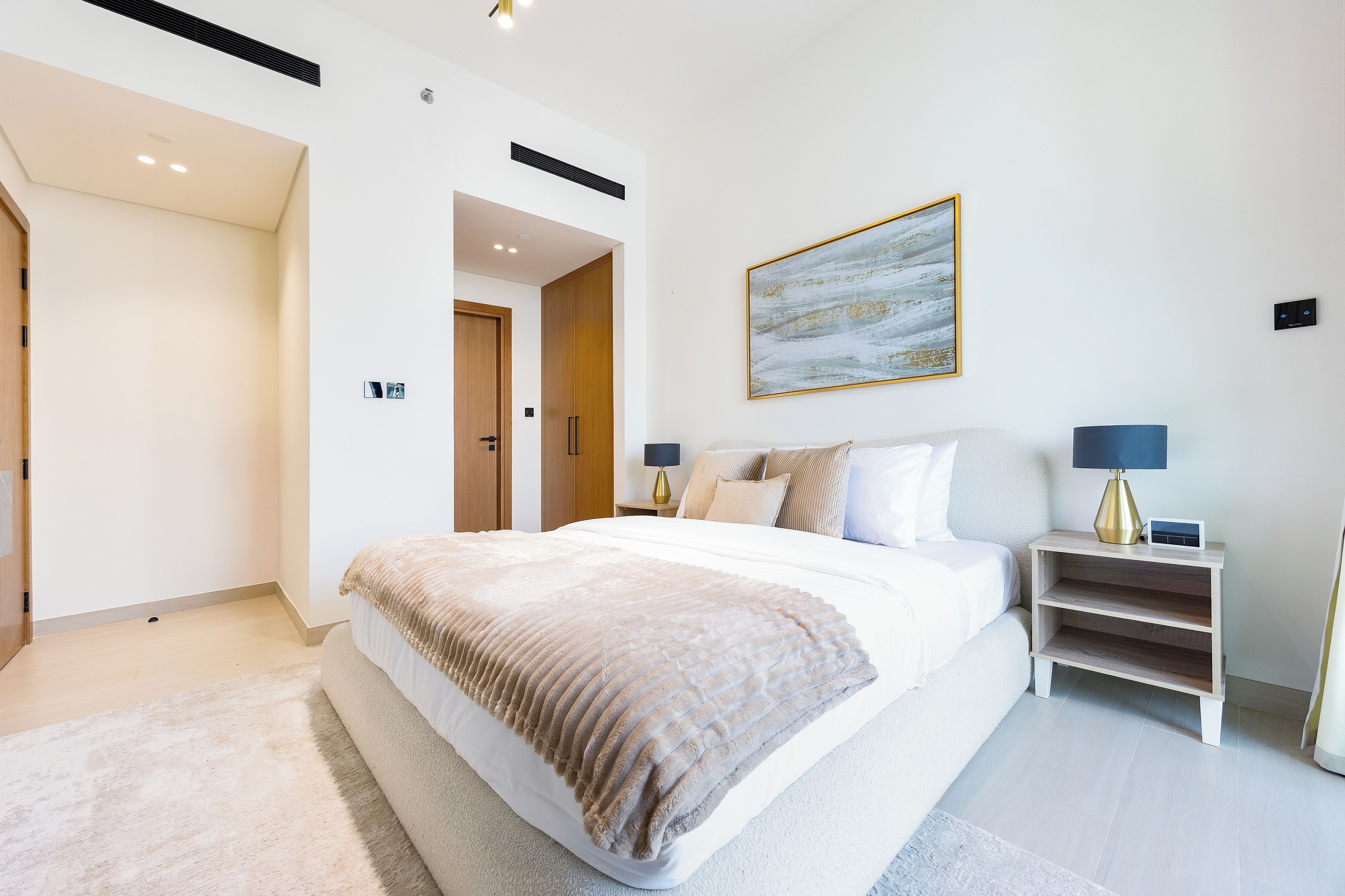 A modern bedroom with a large bed, bedside tables, a painting, and a ceiling vent.