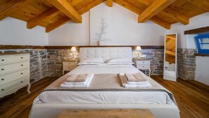Un dormitorio con techo de madera, paredes de piedra, una cama con ropa de cama blanca, dos mesitas de noche con lámparas y un aparador.