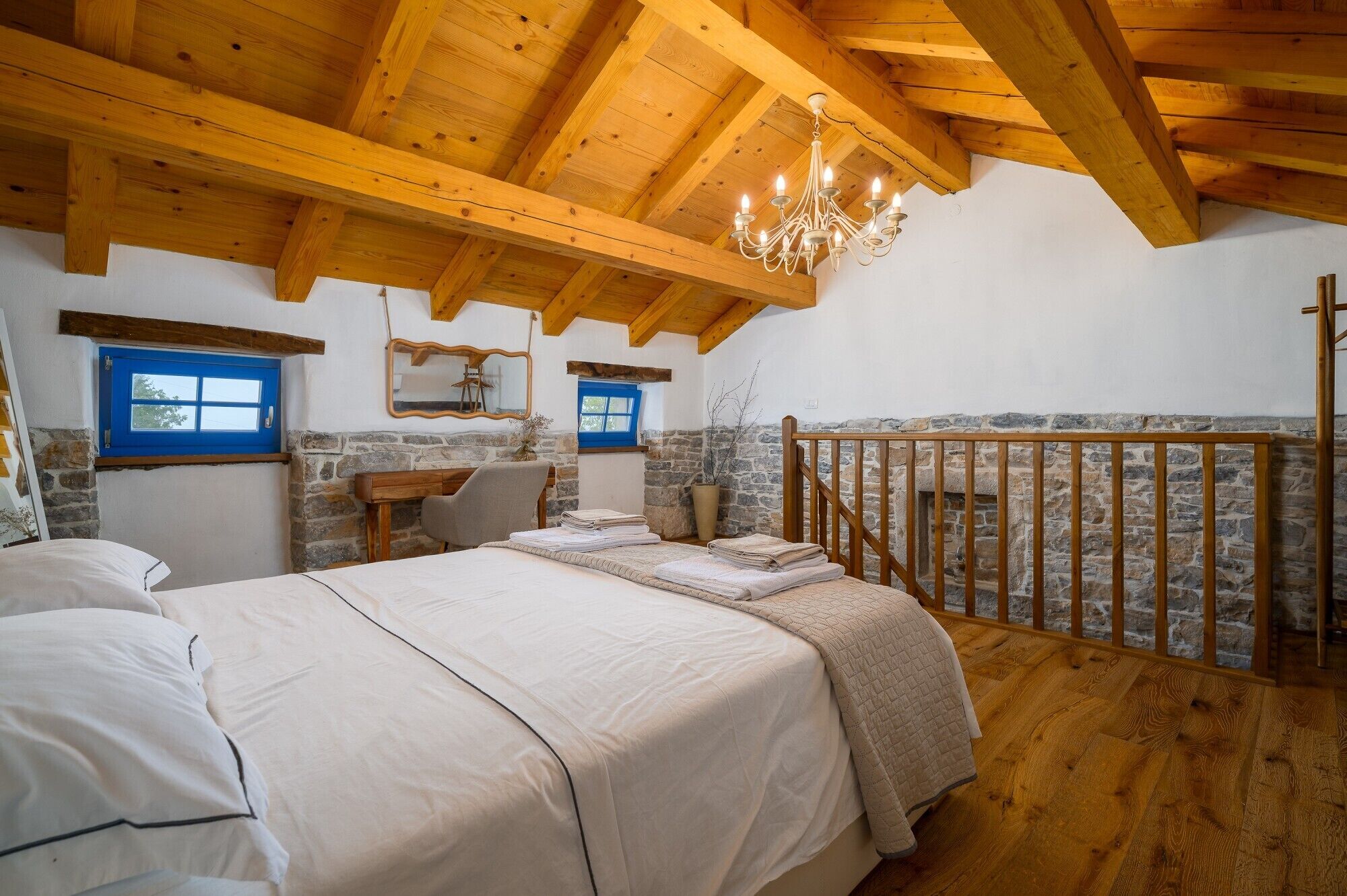 Un dormitorio con una cama grande, un escritorio, una silla, una pared de piedra y barandilla de balcón de madera.