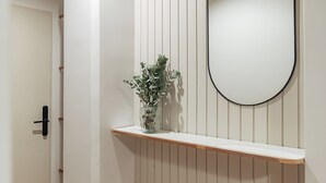 A room with a white door, a wooden shelf with a vase of green plants, and a round mirror on the wall.